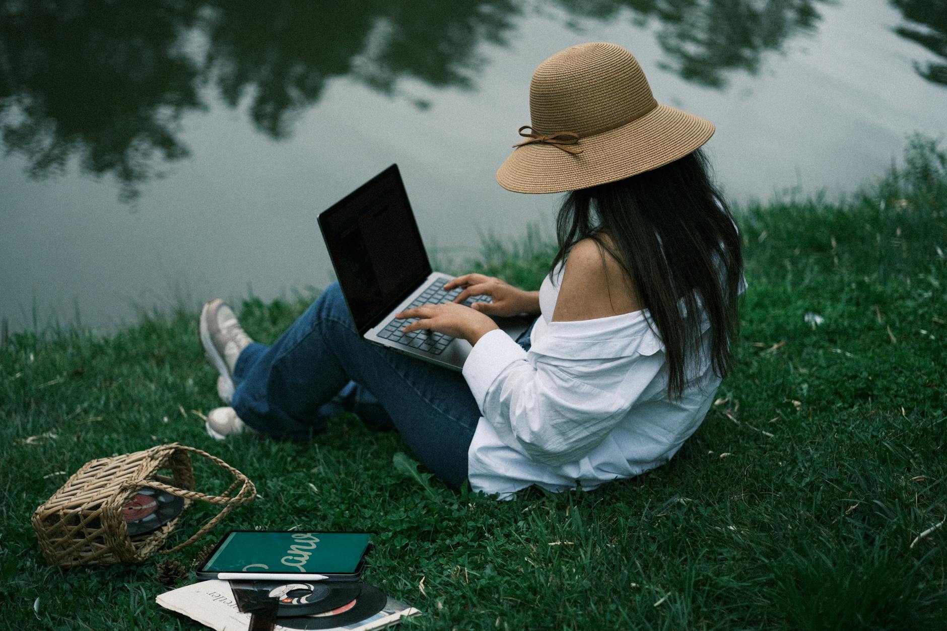 Kobieta w kapeluszu pracuje na laptopie nad spokojnym jeziorem w parku